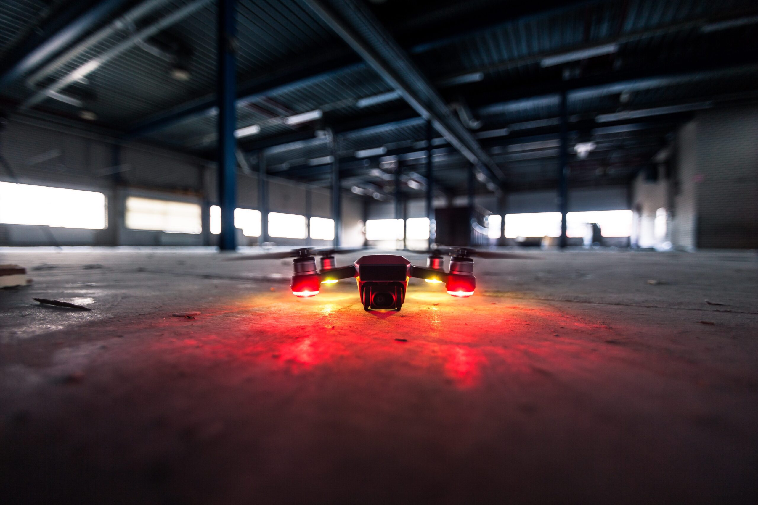 A close-up of a UWB-Based RTLS drone on a concrete floor, in the dark but the drone has red lights that reflects on the floor. The environment looks like an abandoned empty warehouse.